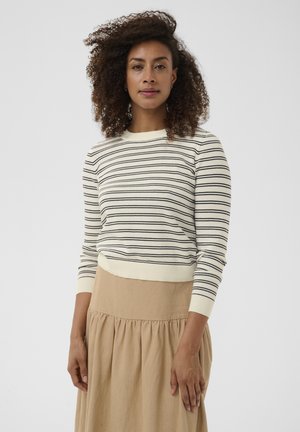 Vrouw met krullend haar, gekleed in een witte trui met zwarte strepen en een beige rok, staand tegen een effen lichte achtergrond.