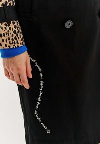 Čierny trenčkot s rukávom v leopardom vzore. Obsahuje gombíkovú sponu a biele textové detaily po boku. Hladká textúra, prispôsobený strih.