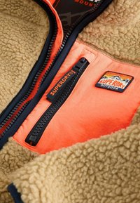 Fleece bunda s hnědým texturovaným tělem a oranžovou podšívkou. Obsahuje černý zip a vyšívanou nášivku "Superdry Outdoor".