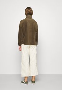 Un pulóver marrón, texturizado, de manga larga y cuello alto, combinado con pantalones anchos color crema y zapatillas verdes.