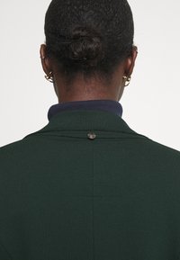 Dark green textured fabric jacket with a navy collar, featuring a small silver logo button at the back neck and visible seam details.