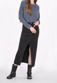 Jupe longue en denim noir avec une fente avant, portée avec un pull en maille bleu. Assortie à des bottines noires ornées d'accents métalliques.