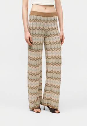 Femme portant un pantalon en maille à jambes larges avec un motif zigzag beige, marron et bleu clair, associé à des talons ouverts et un haut blanc court.