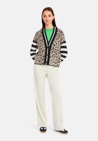 Leopardenmuster-Cardigan mit Schwarz-weiß gestreiften Ärmeln, schwarzer Borte und Knöpfen, kombiniert mit einem grünen Oberteil und weißen weiten Hosen.