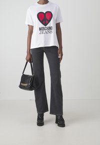 Modelka stojí proti jednoduchému pozadí, oblečená v bílém tričku s červeným symbolem míru a textem "Moschino Jeans", tmavých džínech a černých platformových botách, držící černou kabelku.