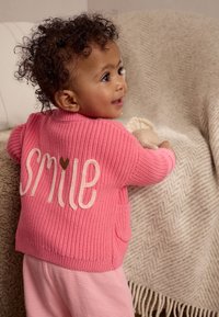 Cardigan rosa lavorato a maglia con la scritta "SMILE" in bianco e un dettaglio a forma di cuore, abbinato a pantaloni rosa a righe. La trama è a coste e confortevole.
