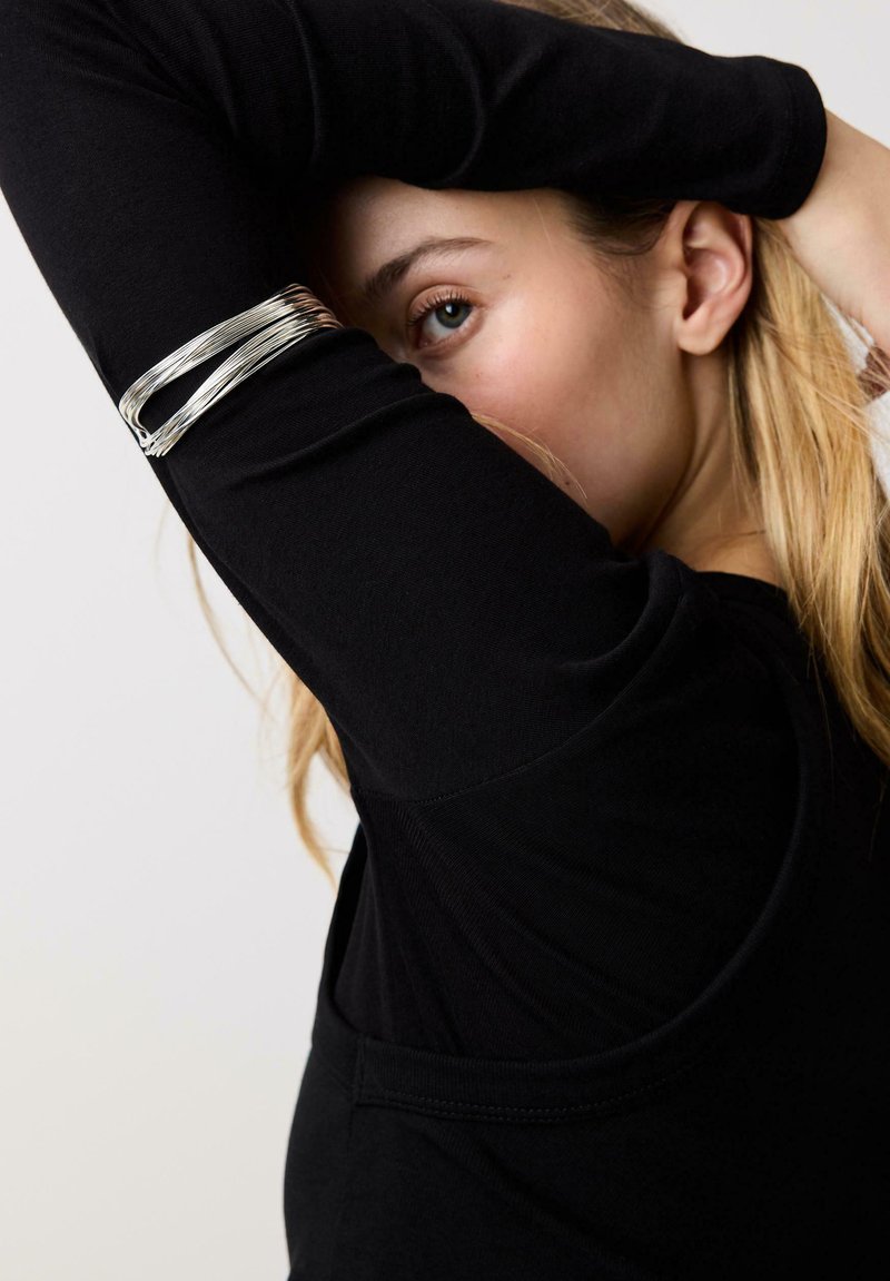 Femme aux cheveux blonds portant un haut noir à manches longues, cachant partiellement son visage avec son bras, et portant un bracelet en argent à plusieurs bandes au poignet.