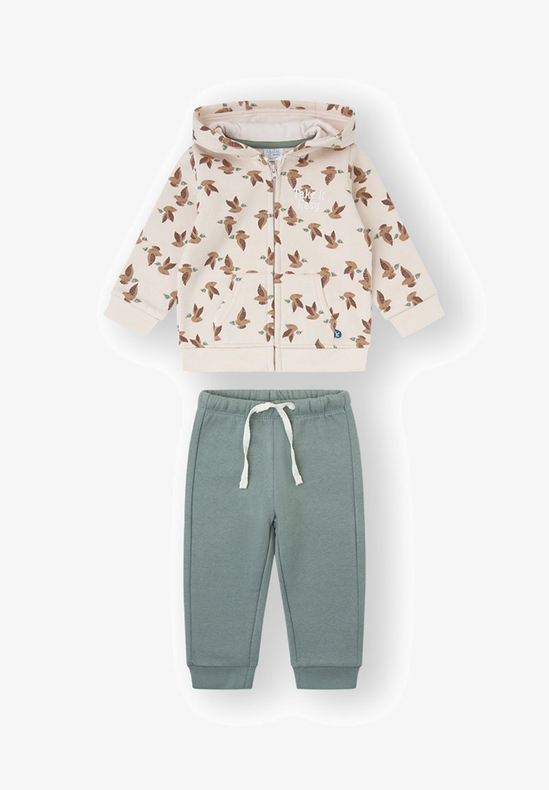Sudadera con capucha y cremallera en color crema con estampado de pájaros, combinada con pantalones deportivos verdes que cuentan con cintura y puños elásticos.