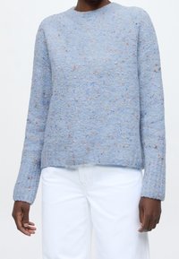 Personne portant un pull bleu clair texturé à manches longues avec des taches multicolores, associé à un pantalon blanc sur fond blanc.