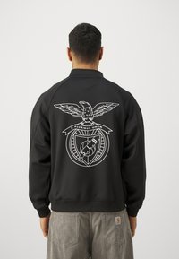 Sort sweatshirt med høj krave, prydet med et hvidt ørn emblemb og tekst på ryggen. Fremstillet af glat materiale med en afslappet pasform.