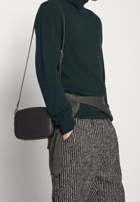 Mörkgrön polotröja med matchande knuten midjedetalj, tillsammans med svarta och vita mönstrade byxor och en liten svart crossbody-väska.