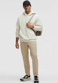 Beige színű kapucnis pulóver, krémszínű, kenguruzsebbel, világos bézs nadrággal és fekete-fehér sportcipőkkel kombinálva. Kiegészítők között szerepel egy fekete sapka és egy bézs táska.