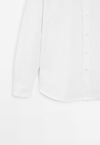 Chemise blanche à manches longues, boutons simples, manchette et ourlet arrondi sur fond uni.