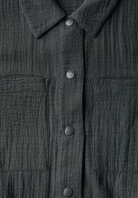 Dunkelgraues Hemd mit Knopfleiste, aus strukturiertem Stoff mit einem dezenten, wellenförmigen Muster und zwei Brusttaschen. Die Vorderseite wird von schwarzen Plastikknöpfen geschlossen.