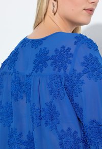 Blå gennemsigtig top med indviklet blomstret broderi i matchende tone, med en afslappet pasform og en rund halsudskæring. Minimal hardware-detaljer.