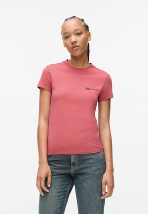 Støvet rosa t-skjorte med korte ermer laget av bomull, med rund hals og svart logotekst på venstre side. Brukt med blå jeans.