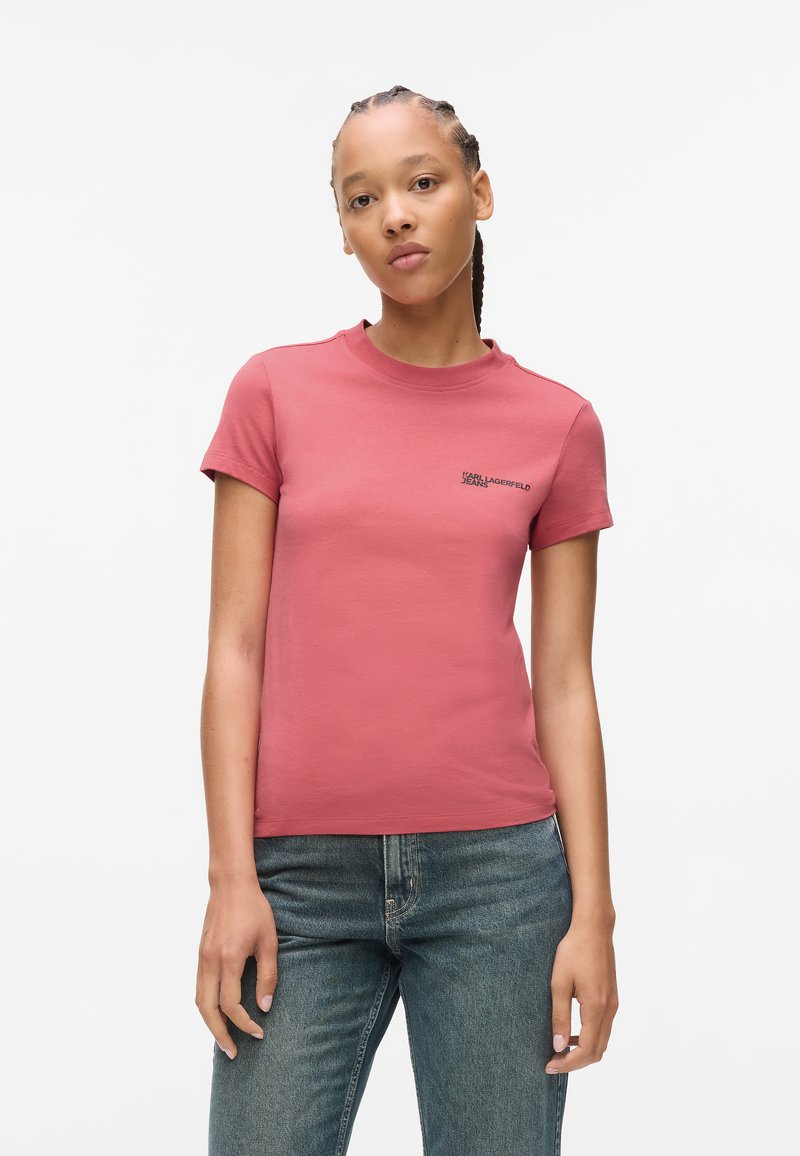 Støvet rosa t-skjorte med korte ermer laget av bomull, med rund hals og svart logotekst på venstre side. Brukt med blå jeans.