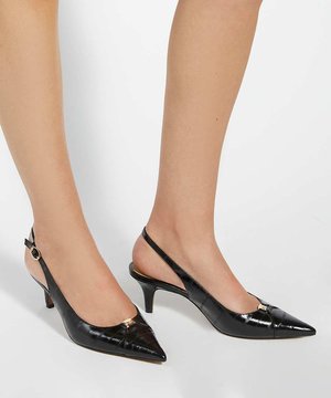 Jambes portant des escarpins slingback en cuir verni noir à petit talon kitten, bouts pointus et petites décorations dorées sur fond blanc.