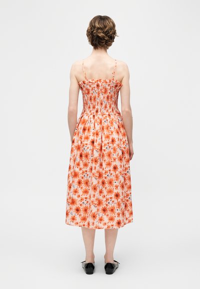Γυναίκα με κοντά μαλλιά που φορά ένα πορτοκαλί φόρεμα με φλοράλ μοτίβο, χωρίς μανίκια, μήκους μέχρι τα μέσα της γάμπας, με σμιλευμένο μπροστινό μέρος, φωτογραφημένη από πίσω σε μονόχρωμο φωτεινό φόντο.