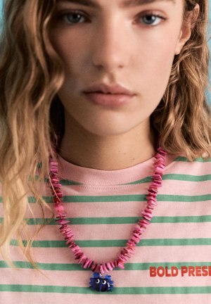 Persoană tânără cu păr blond ondulat, purtând un tricou cu dungi roz și verzi și un colier roz cu mărgele cu pandantiv albastru în formă de crab.