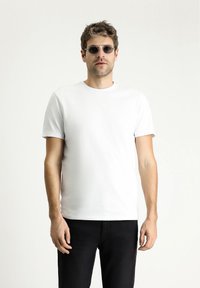 Camiseta blanca de algodón con mangas cortas, cuello redondo y una textura suave; combinada con jeans negros y gafas de sol redondas oscuras.