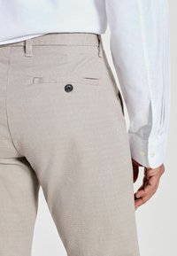 Pantalons beiges clairs texturés avec un motif subtil, deux poches arrière et un détail de bouton noir, associés à une chemise blanche à manches longues.