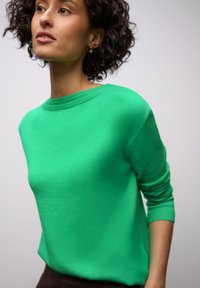 Pull vert à col rond, en tissu tricoté texturé, avec manches longues. Des coutures près des épaules ajoutent un intérêt subtil.