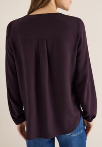 Bordowa bluzka z długim rękawem, okrągłym dekoltem oraz plisą z tyłu, z elastycznymi mankietami, w połączeniu z niebieskimi dżinsami.