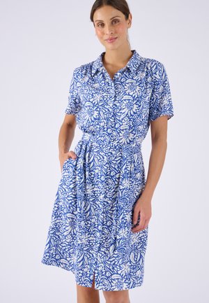 Mujer que lleva un vestido corto de flores azul y blanco con cuello y botones al frente, de pie con una mano en el bolsillo.