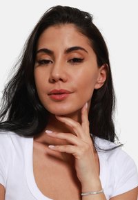 Donna con lunghi capelli neri che si tocca il collo, indossando una maglietta bianca aderente. I dettagli notevoli includono unghie di un rosa chiaro e un braccialetto d'argento.