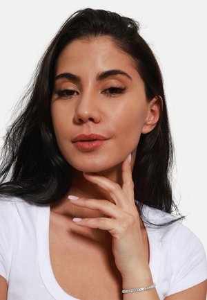 Donna con lunghi capelli neri che si tocca il collo, indossando una maglietta bianca aderente. I dettagli notevoli includono unghie di un rosa chiaro e un braccialetto d'argento.