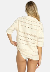 Suéter de punto oversize color crema con mangas cortas, que presenta un tejido abierto a rayas y un cuello acanalado. Textura arrugada con aberturas laterales.