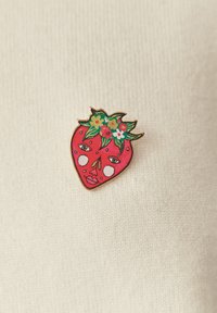 Broche en émail en forme de fraise avec un visage ; présente une couleur rouge, des feuilles vertes, des accents floraux et des yeux cartoon avec des détails blancs.