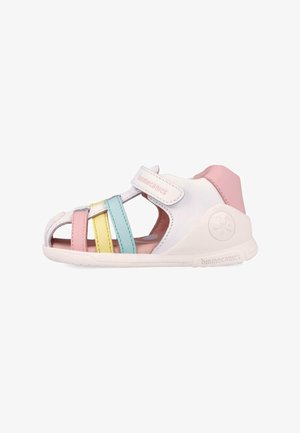 Sandalia blanca para niño pequeño con tacón rosa, correa ajustable y tiras en tonos pastel rosa, amarillo y azul en la suela blanca.