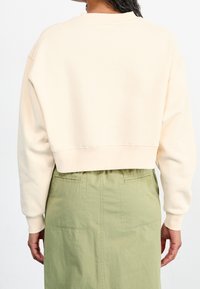 Persona che indossa una felpa accorciata color crema e una gonna a vita alta verde chiaro, vista da dietro su uno sfondo bianco.