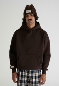 Hombre con sudadera con capucha marrón oscuro y gorro a juego con etiqueta, combinado con pantalones a cuadros en blanco y negro, de pie frente a un fondo liso.