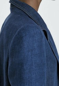 Tummansinisestä denim-takista, jossa on klassinen kaulus, näkyvät saumat ja muotoillut hartiat. Sileä tekstuuri ja istuva siluetti.