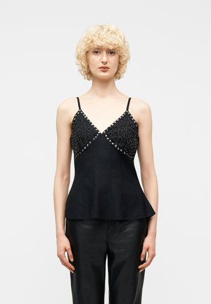 Lyshåret person iført en sort ærmeløs top med sølvnitter og mønstre kombineret med sorte bukser, stående mod en ensfarvet baggrund.