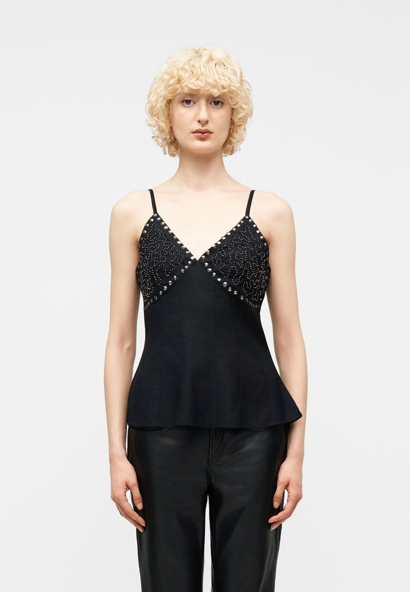 Ξανθό άτομο που φοράει μαύρο αμάνικο τοπ με ασημένιες τρουκς και σχέδια, συνδυασμένο με μαύρο παντελόνι, στέκεται μπροστά σε απλό φόντο.