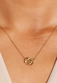 Gouden ketting met een getextureerde knoop hanger aan een delicate ketting, rustend tegen een gladde huidskleurige achtergrond.