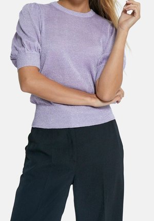 Vrouw draagt een lichtpaars glanzend kort mouw topje met pofmouwen en zwarte hoog getailleerde broek, armen gekruist op de taille.