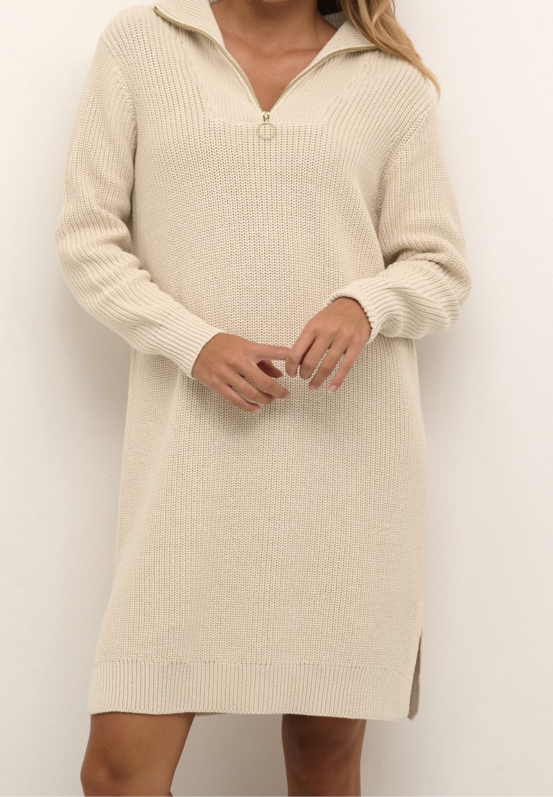 Femme portant une robe pull en tricot beige à manches longues avec une fermeture éclair frontale et un col, se tenant devant un fond clair uni.
