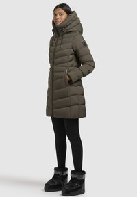 Khaki puffer coat with a high collar, zip closure, and side pockets. Paired with black leggings and boots, featuring a cushioned design.