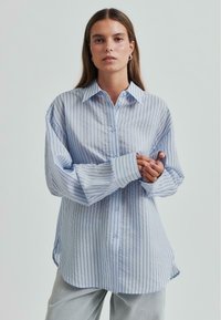 Cămașă butonată de culoare albastru deschis, cu dungi verticale, mâneci lungi și guler. Realizată dintr-un material moale, cu o croială lejeră și un tiv curbat.