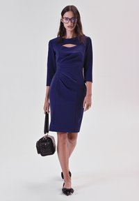 Navyblaues, tailliertes Kleid mit Dreiviertelärmeln, das über einen Ausschnitt am Ausschnitt und geraffte Seitennaht verfügt. Kombiniert mit einer schwarzen Handtasche.