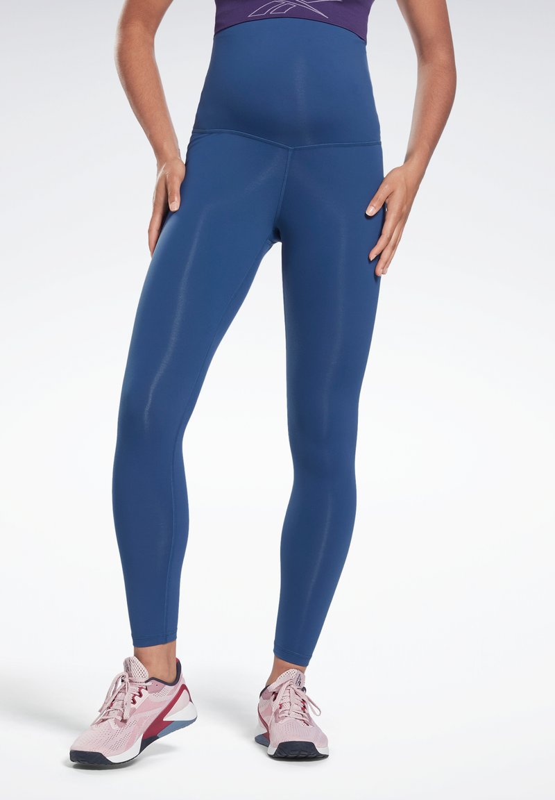 Leggings de cintura alta, justos ao corpo, em tecido azul escuro sólido. Textura suave com um acabamento brilhante. Combinados com sapatilhas de treino rosas.