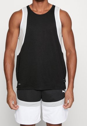 Muskularna osoba ubrana w czarno-biały sportowy tank top bez rękawów i czarno-białe siateczkowe szorty sportowe na jednolitym tle.