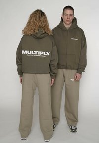 Sweat à capuche zippé vert olive et pantalon large beige. Le sweat à capuche présente un grand logo blanc "MULTIPLY" au dos, une texture douce et une coupe décontractée.