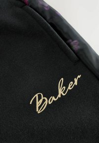 Melna sporta jaka ar gludu tekstūru, kas rotāta ar zelta izšuvumu "Baker" un krāsainu ziedu motīvu iekšējā apkaklīte.
