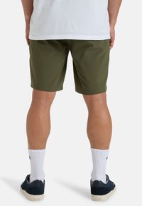 Pantaloni corti verdi oliva con una texture liscia, di lunghezza fino a metà coscia e un design semplice; indossati con calze sportive bianche e sneakers scure.